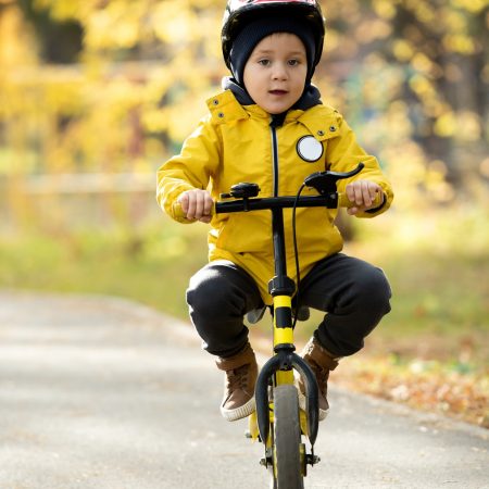 Adorable little boy in casualwear and protective helmet sitting on balance bike in front of camera on asphalt road against natural environment