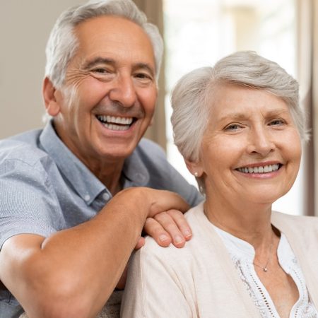 Portrait of happy healthy senior couple at home. Romantic old couple sitting together on sofa while looking at camera. Cheerful elderly wife and husband enjoying life after retirement.