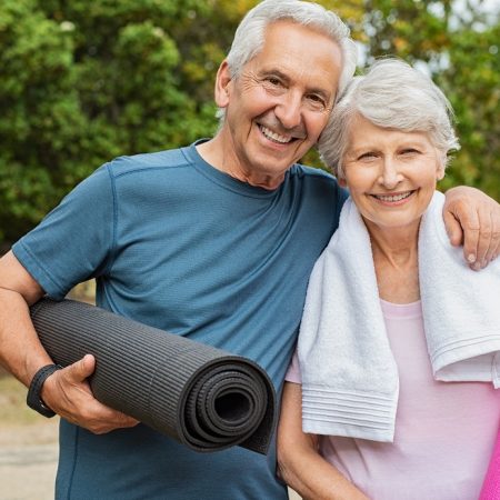 Lovely senior man and old woman holding yoga mat for exercising outdoor. Aged husband and wife ready for yoga session at park. Active healthy retired couple looking at camera before a postural session.