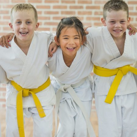 A group of elementary age children are taking a martial arts class. They are standing together in a row and are smiling while looking at the camera.