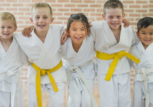 A group of elementary age children are taking a martial arts class. They are standing together in a row and are smiling while looking at the camera.