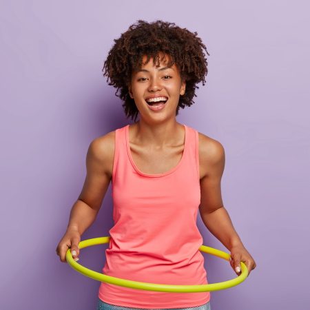 Photo of optimistic dark skinned woman rotates hule hoop, aims to have perfect waist, smiles broadly, likes sport and gymnastics, has fun during exercising, wears casual outfit. Loosing weight concept