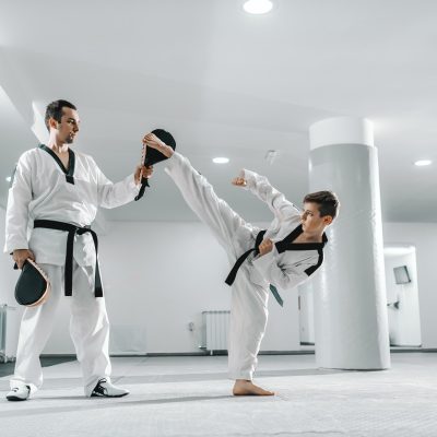 Young Caucasian boy in dobok kicking barefoot while trainer holding kick target. Taekwondo training concept.