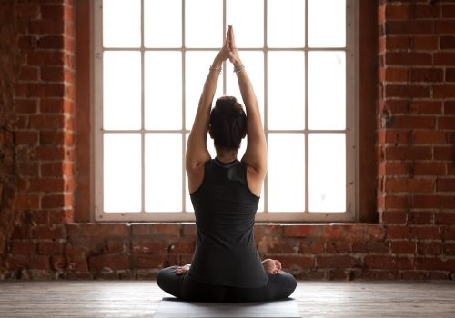 Young sporty woman practicing yoga, doing Sukhasana exercise, Easy Seat pose, working out, wearing sportswear, black pants and top, indoor full length, yoga studio window, rear view